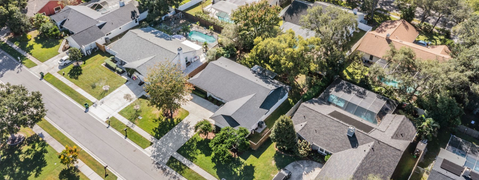Aerial view of suburban homes with gray roofs, green lawns, and tree-lined streets.