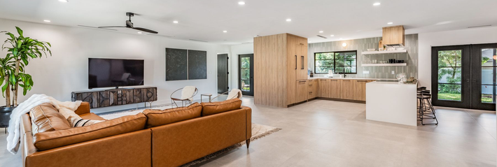Bright open-plan living room with brown sectional sofa, TV, wood cabinets, kitchen, and glass doors