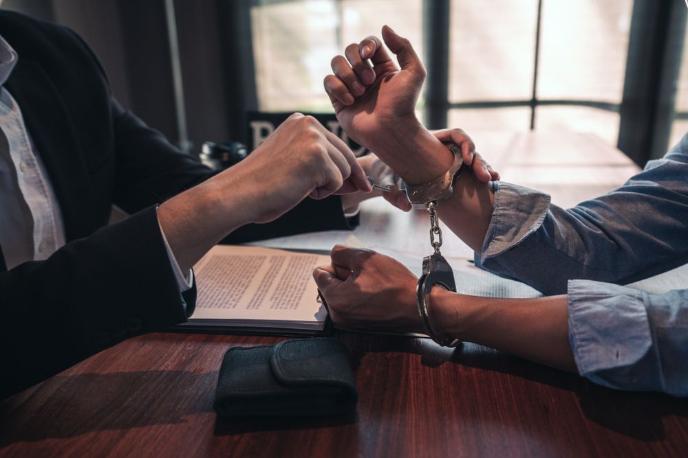 Person in handcuffs at a table, another person placing them on.