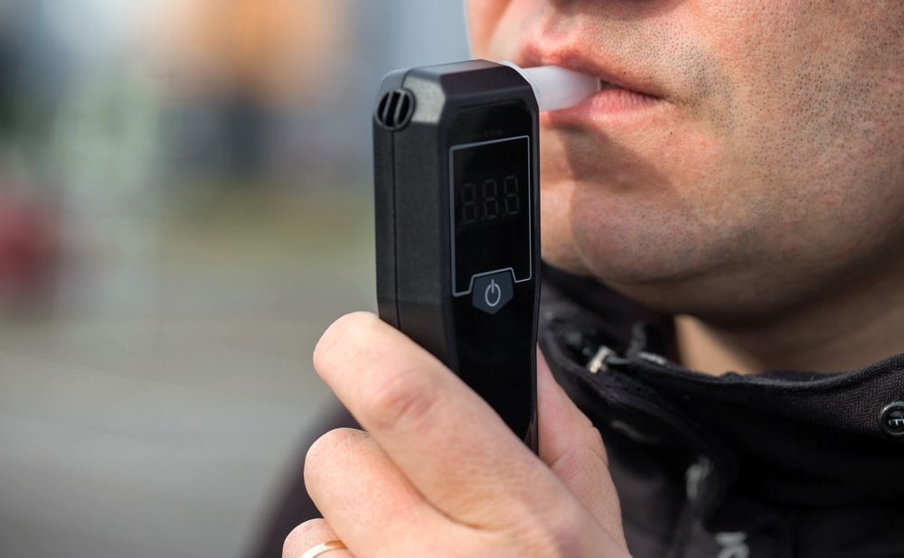 Man blowing into a black breathalyzer device outdoors, testing blood alcohol content.