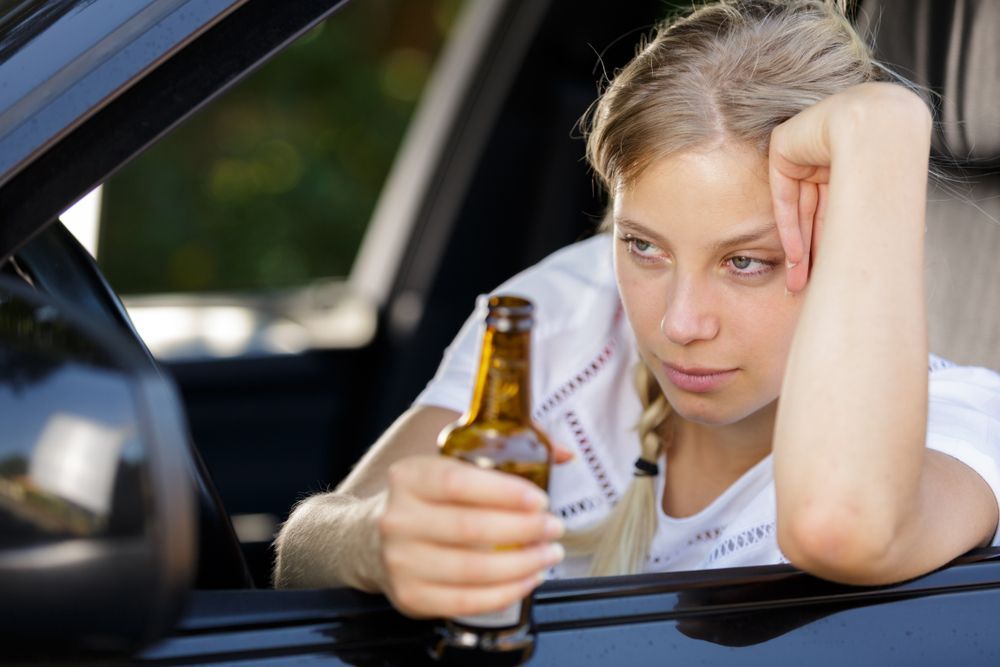Woman in car holding beer bottle, looking pensive.