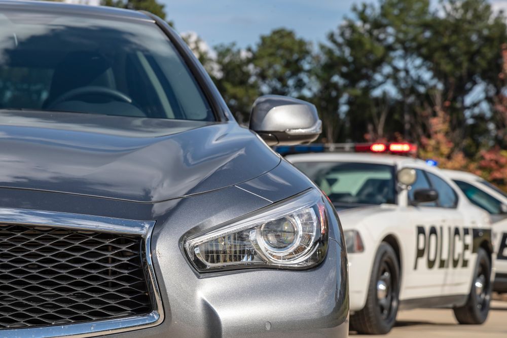Gray car with a focus on its front, and a police car with flashing lights blurred in the background.