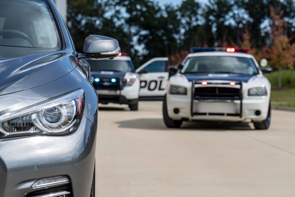 Gray car in foreground, police cars with flashing lights in background, on a road.