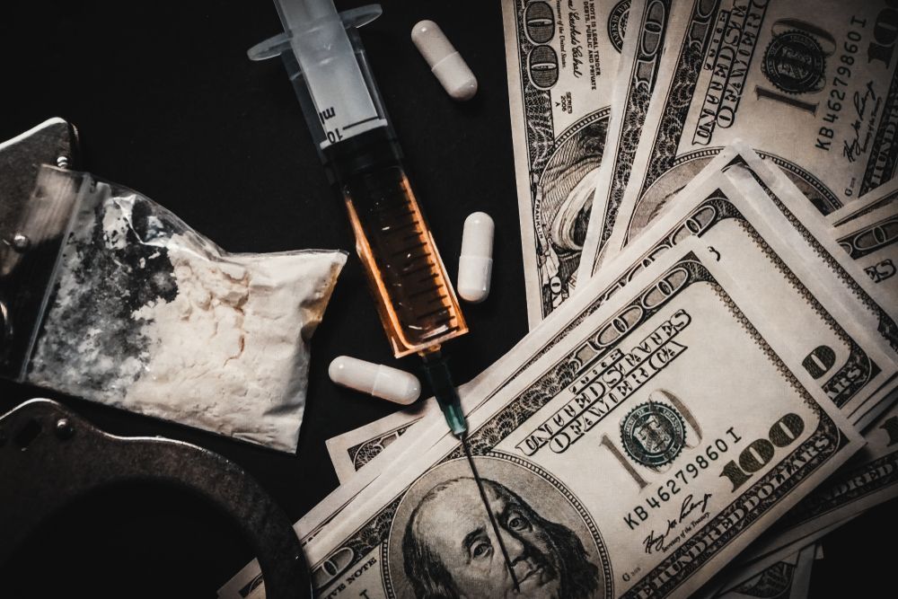 Handcuffs, cash, bag of powder, syringe with liquid, and pills on a dark surface.