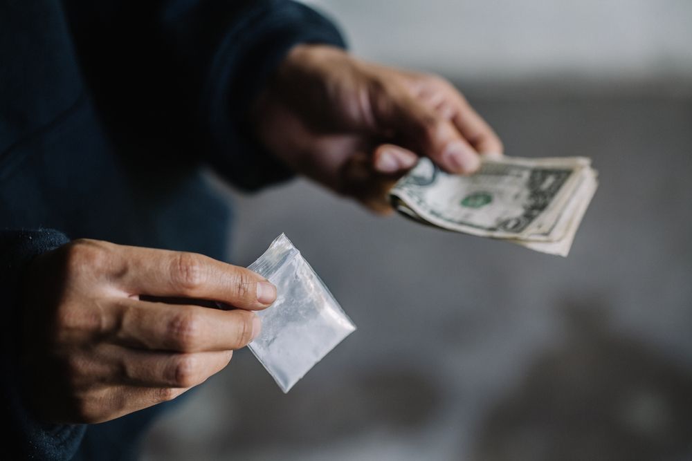 Person's hands holding a small bag of white powder and US dollar bills, likely a drug deal.