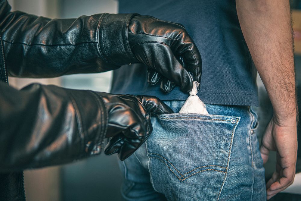 Black-gloved hands planting a small bag of white powder into the back pocket of a person's jeans.