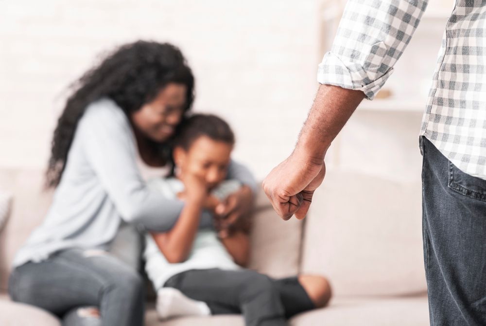 Man's clenched fist in foreground; blurred woman comforts crying child on sofa.