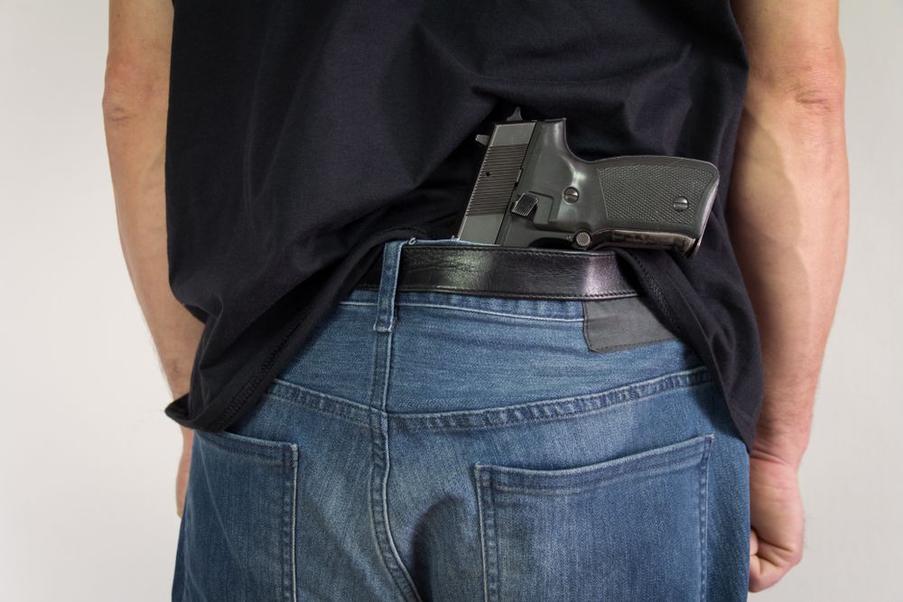 Person's back, a handgun partially visible, tucked in the waistband of jeans, under a black shirt.