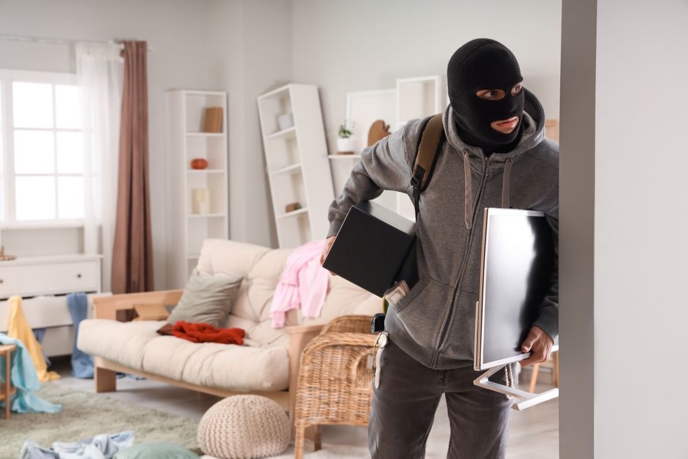 A masked person carrying a computer monitor and laptop exits a messy room.