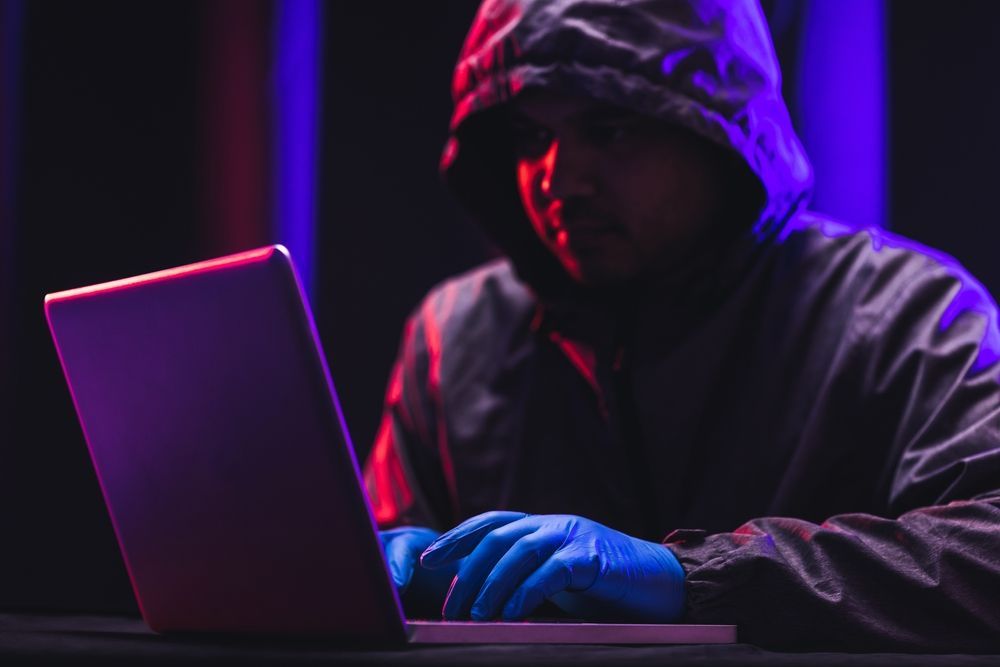 Person in a hooded sweatshirt and gloves using a laptop in a dark room illuminated with red and blue light.