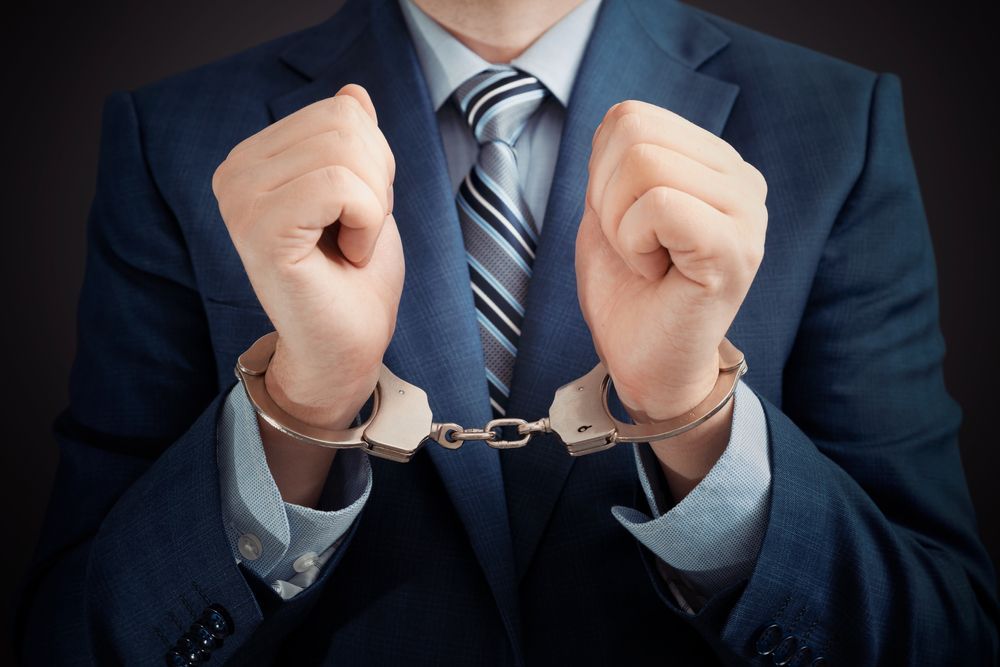 Person in a suit with hands cuffed in front of them, centered against a dark background.