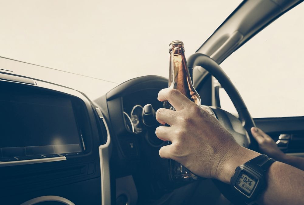 Person holding a beer bottle in a car while gripping the steering wheel.
