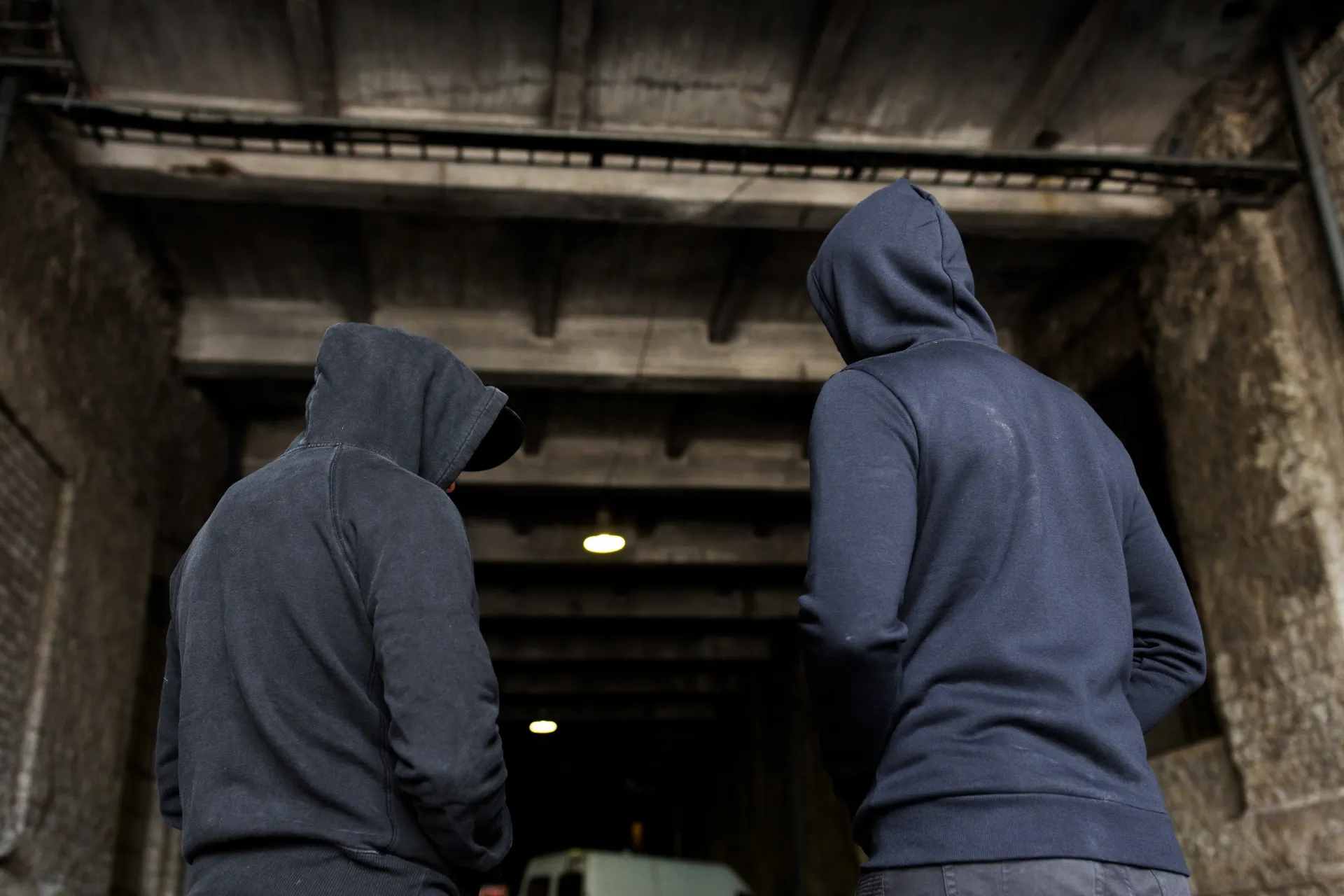 Two people in hooded sweatshirts stand in a dark tunnel, facing away from the viewer.