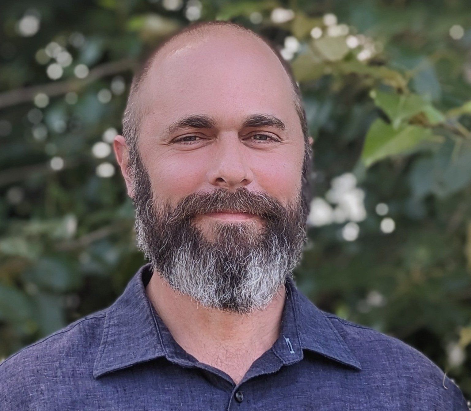 Man with a beard and short hair smiles, wearing a blue shirt, outdoors in front of leafy green background.