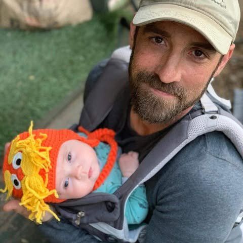 Man holding baby in a carrier, both looking at the camera. Baby wears a monster hat.