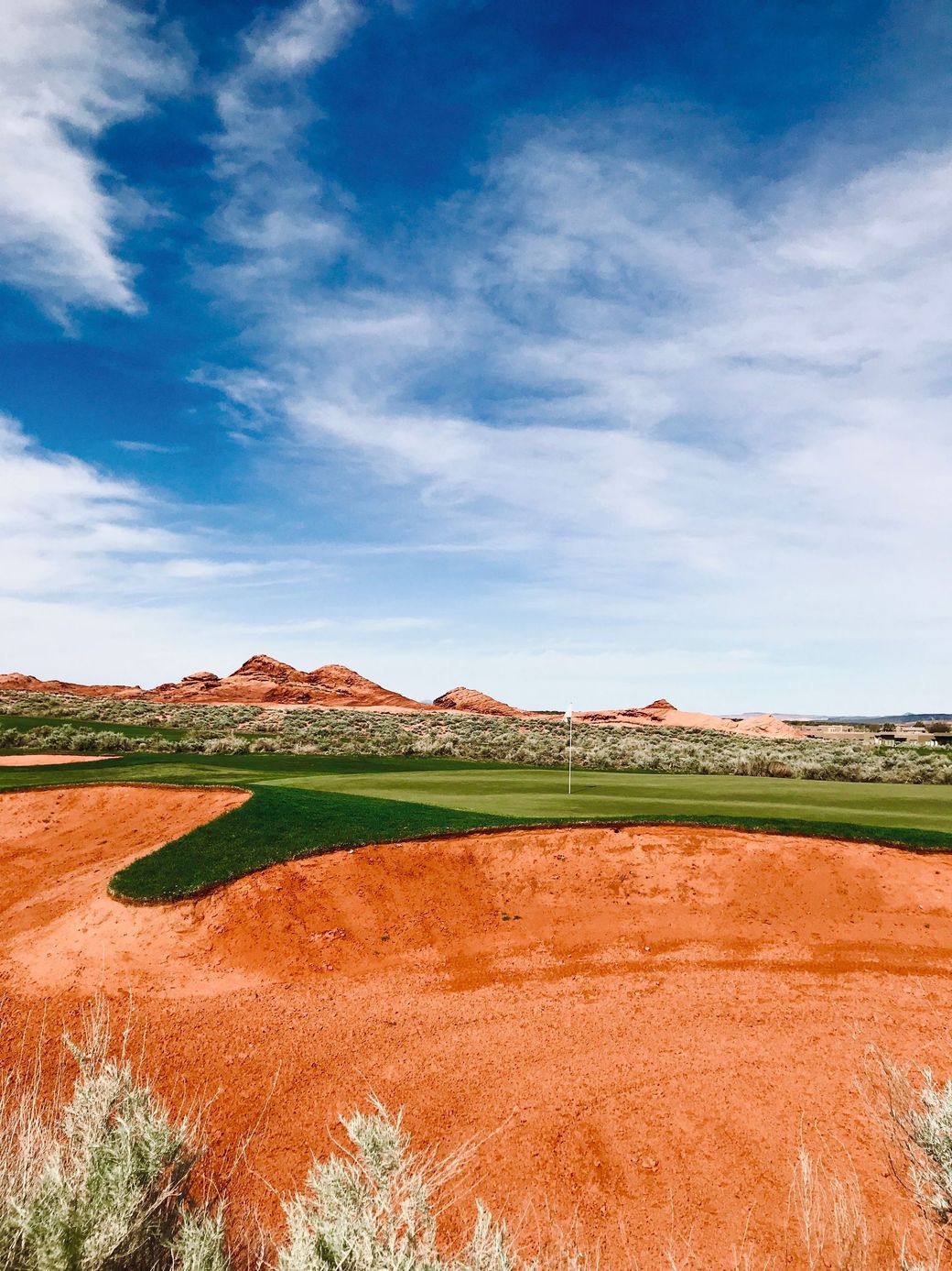 Red desert landscape with green golf course under a blue sky with clouds.