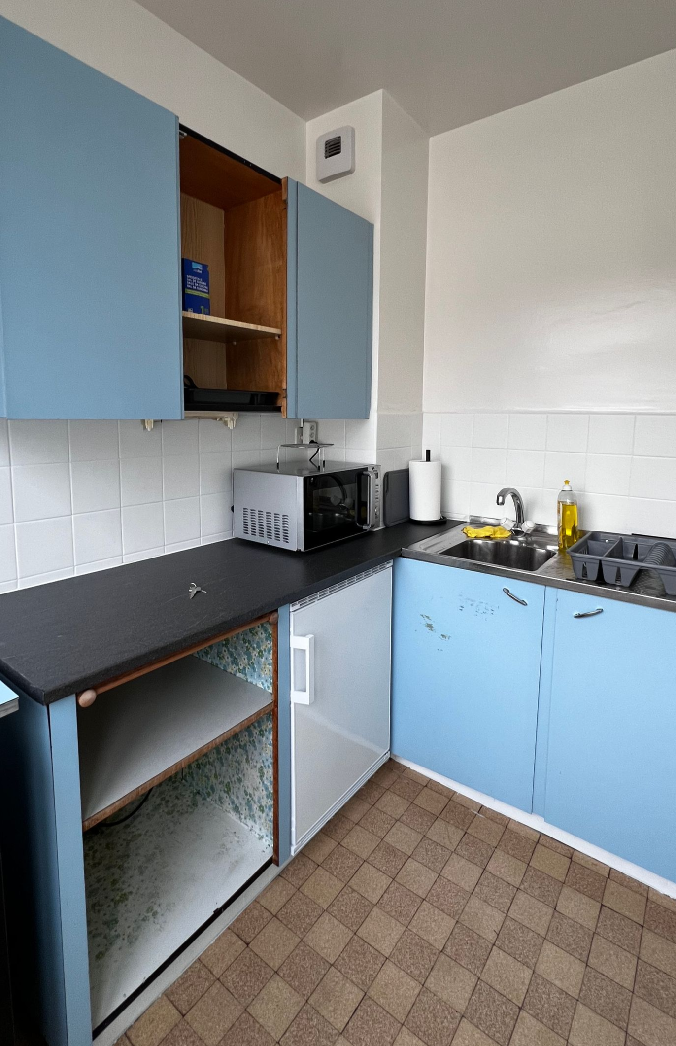 Cuisine compacte avec placards bleu clair, plan de travail noir, cuisinière à gaz et carrelage à motifs au sol.