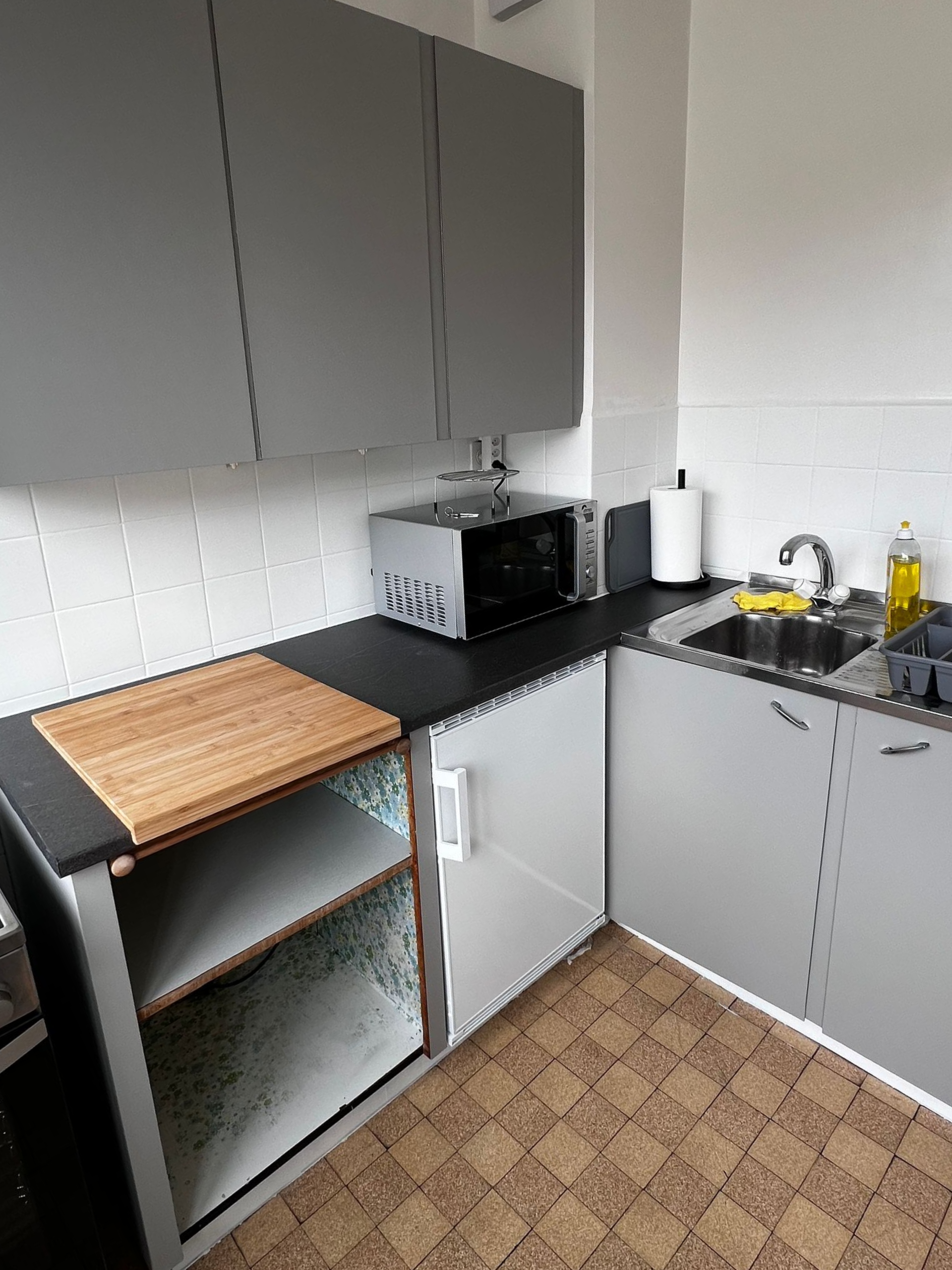Coin cuisine compact avec placards gris, plan de travail noir, micro-ondes, évier et étagères ouvertes.