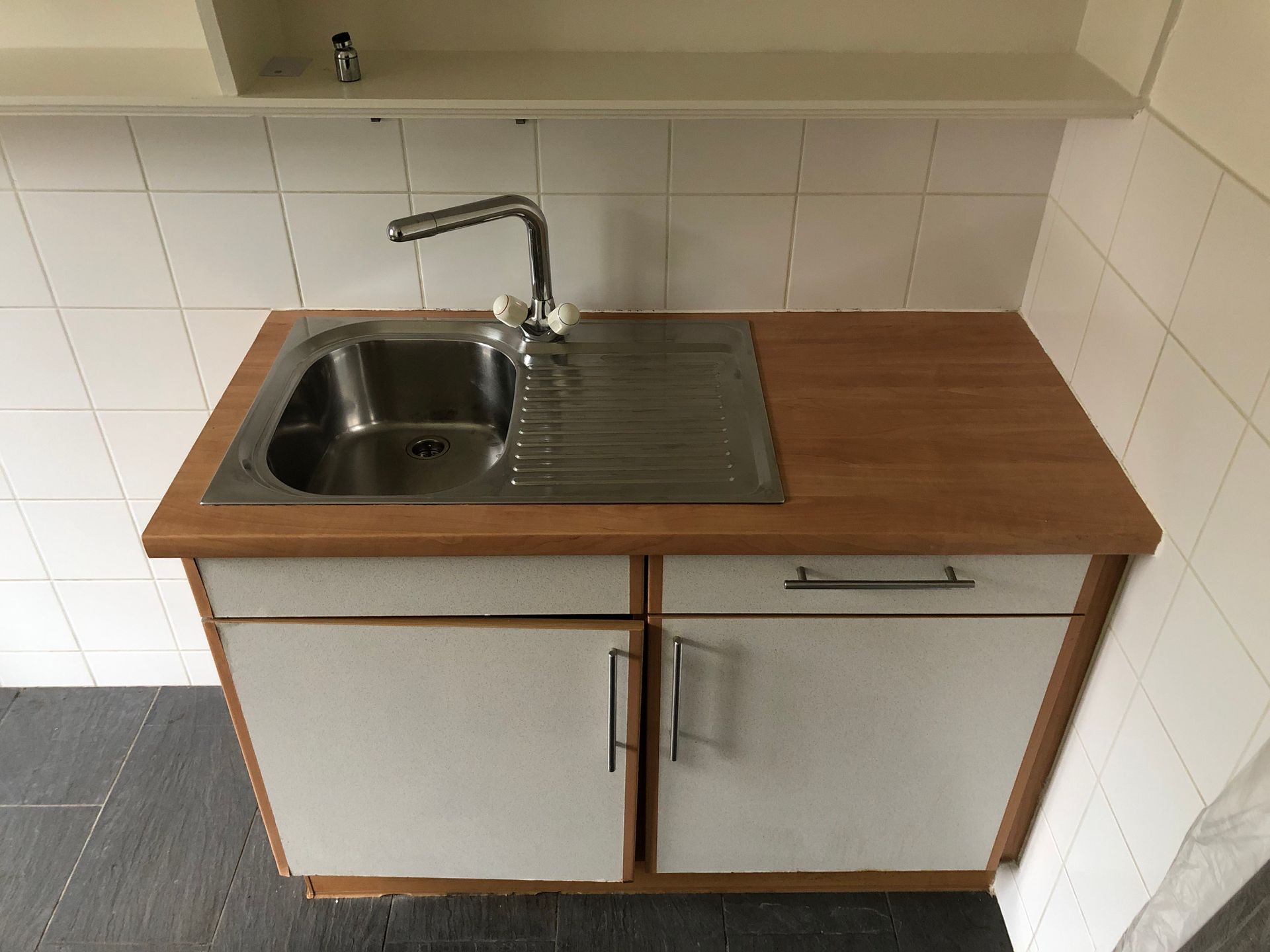 Petit évier de cuisine avec plan de travail, placards blancs et murs carrelés sous une étagère.