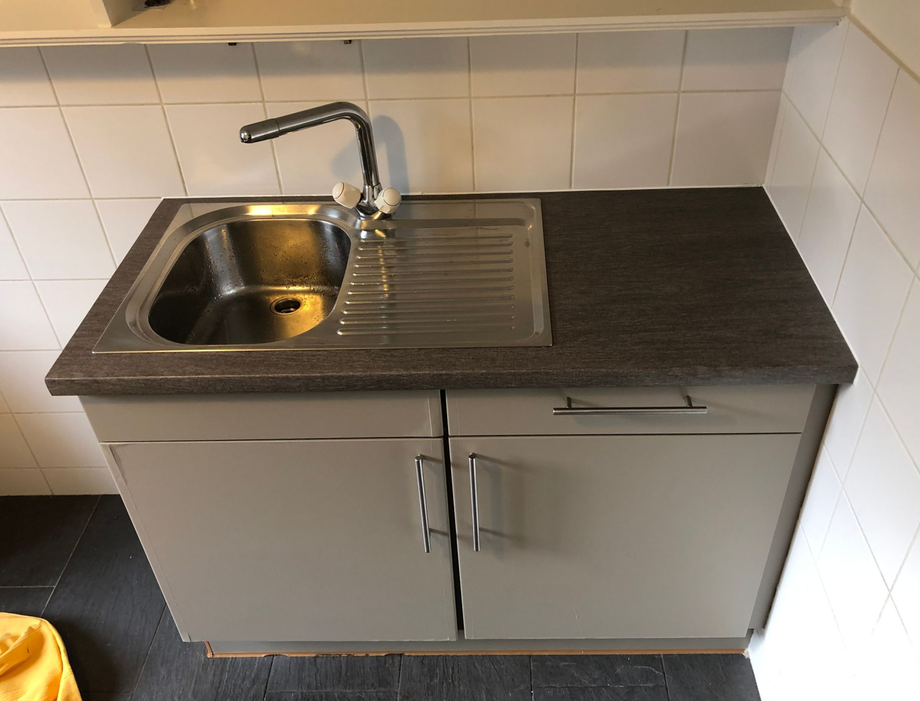 Évier de cuisine compact avec robinet en acier inoxydable et meuble gris sous un plan de travail foncé.