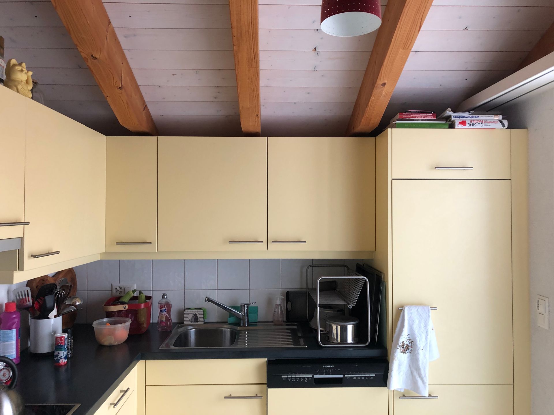 Petite cuisine avec placards couleur crème, plans de travail noirs et poutres apparentes au plafond.