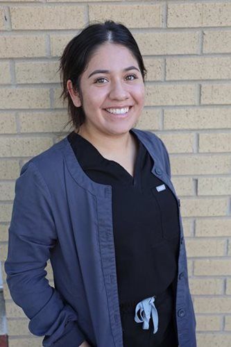 Woman in scrubs and cardigan smiling in front of a brick wall.