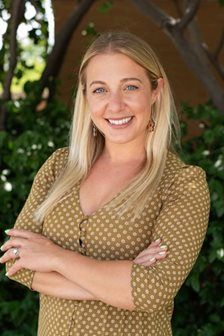 Blonde woman with arms crossed, smiling, wearing a button-down patterned shirt, outdoors.