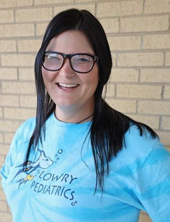 Person wearing glasses and a light blue Lowry Pediatrics t-shirt smiles at the camera.