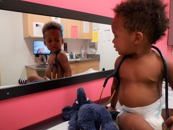 Young child with stethoscope around neck looks at self in mirror in doctor's office.