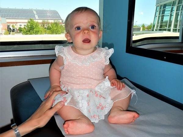Baby sitting on examination table, wearing a pink dress, looking up with mouth open. A hand is reaching out.