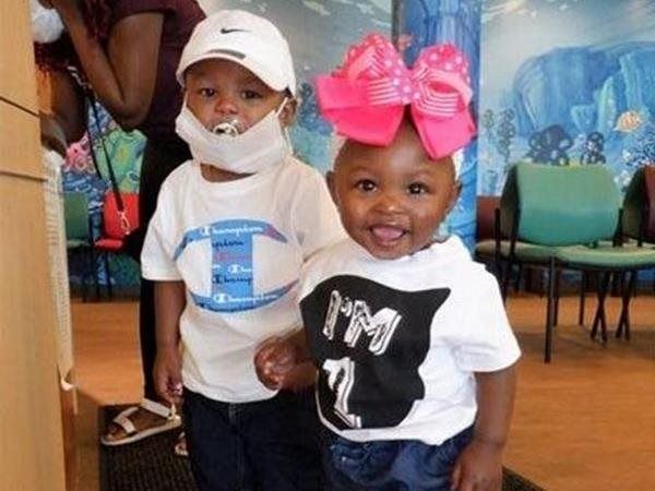 Two toddlers, one with a pacifier and hat, the other with a large pink bow, smiling, holding hands.