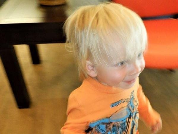 Blond-haired toddler in orange shirt smiles, indoors.