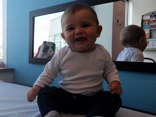 Smiling baby in white shirt and dark pants sitting indoors, reflected in a mirror.