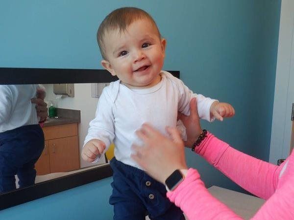 Baby smiles while being held, wearing white long-sleeved shirt and blue pants. Pink-sleeved arm helps. Turquoise wall behind.