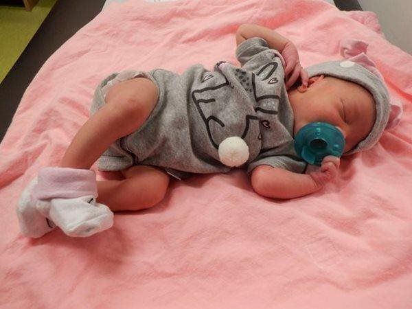 Newborn baby in gray outfit with pacifier and cap, lying on pink blanket.