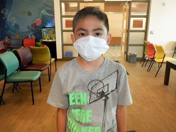 Boy in gray t-shirt and surgical mask standing indoors, wooden floor, chairs in the background.
