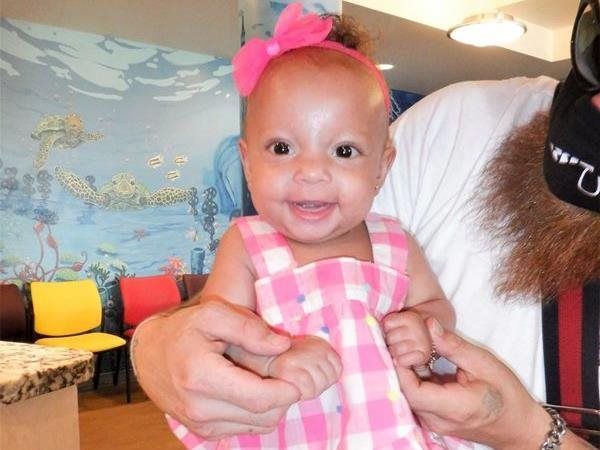 Smiling baby in pink gingham dress, pink bow, being held by a person.
