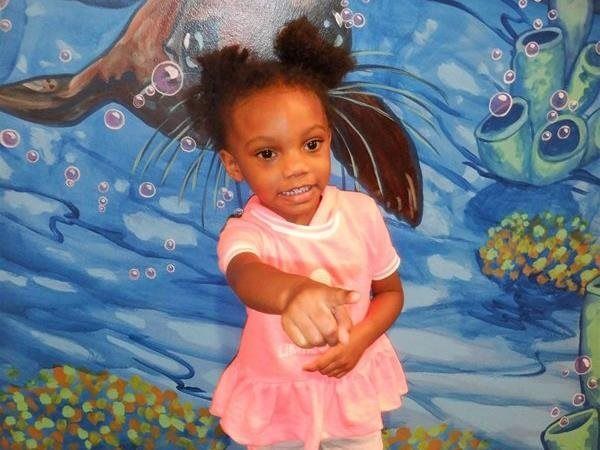 Girl in pink shirt pointing forward, smiling in front of a blue underwater-themed mural.