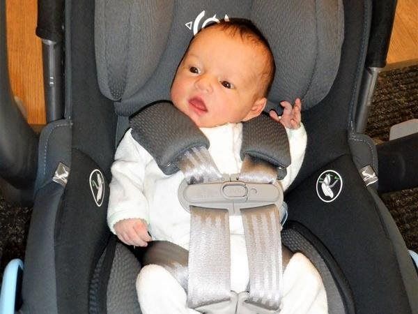 Newborn in car seat, secured with gray harness and shoulder pads, looking up with open mouth.