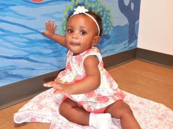 A baby sits near a blue mural, wearing a floral outfit and white headband, with a matching blanket.