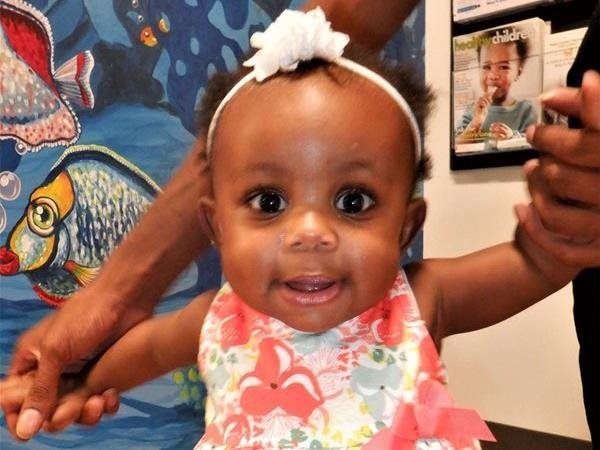 Smiling baby in floral dress and white headband, held by hands, in front of a blue fish mural.