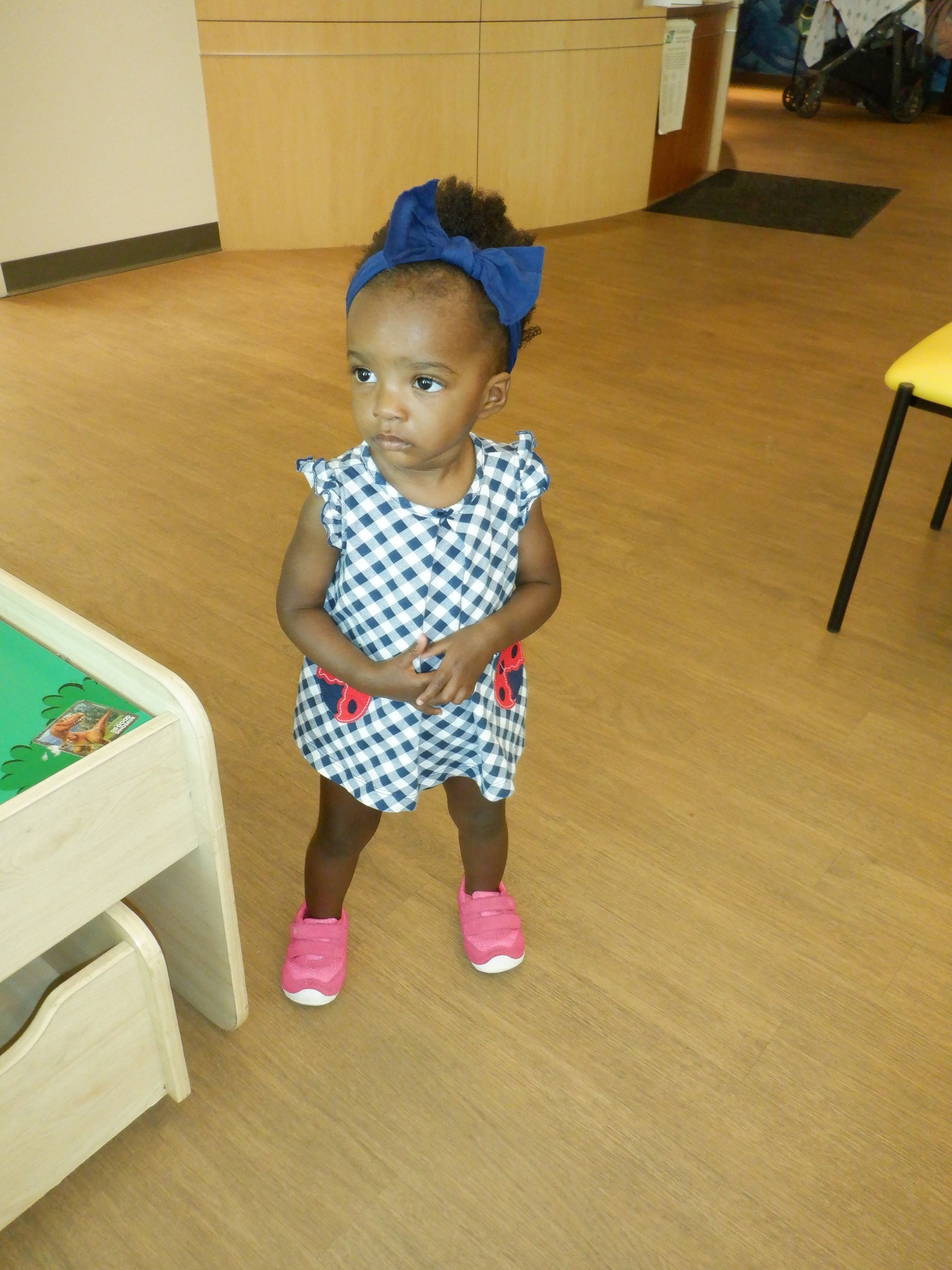 Child in blue and white checkered dress, pink shoes, and blue headband stands in a room.