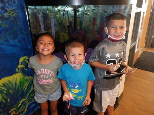 Three children smiling in front of an aquarium. One child has a neck brace. Bright, colorful setting.