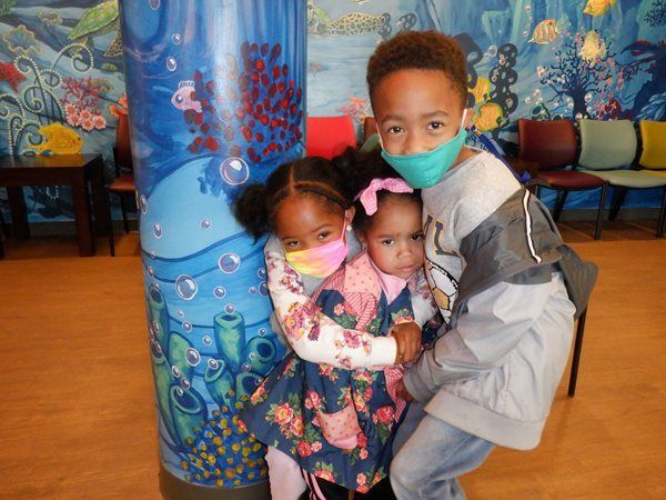 Three children wearing masks, huddled together by a blue pillar with underwater artwork; a waiting room.