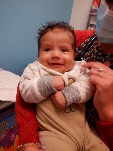 Smiling baby in a beige onesie, held by a person with a red shirt and a medical mask.