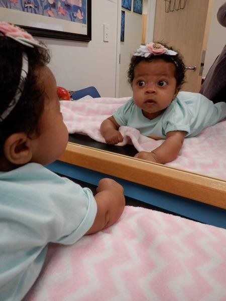 Baby in a light blue onesie, looking surprised at its reflection in a mirror. Pink blanket.