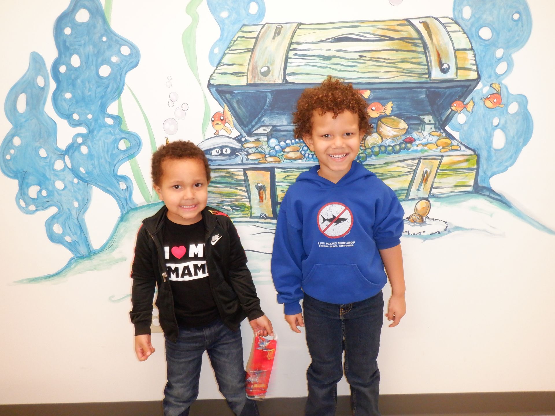 Two children stand in front of a treasure chest mural. One wears a black jacket, the other a blue hoodie.
