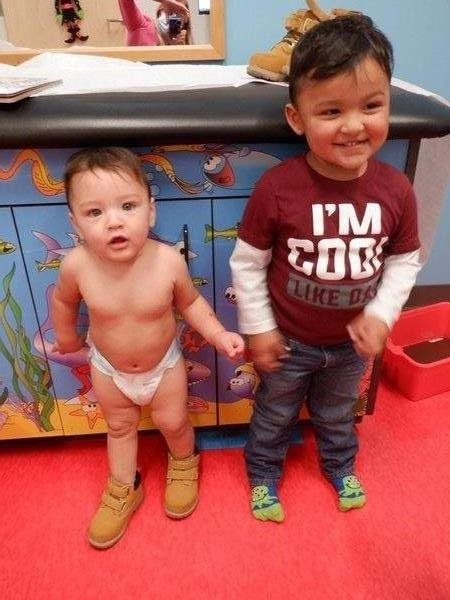 Two young children, one in a diaper, the other in a shirt and jeans, wearing brown boots. Standing in a room with blue cabinets.