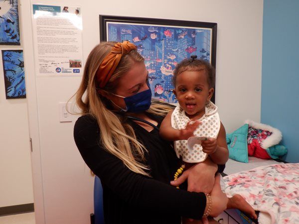 Woman in mask holds smiling baby in a medical room, artwork on wall.