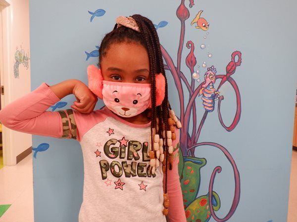 Girl in pink mask and earmuffs flexing arm in front of an aquatic-themed mural.